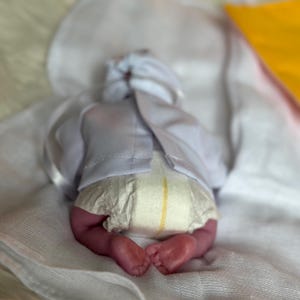 K&ouml;nnte beinhalten: Eine realistische Neugeborenenpuppe, die auf dem Bauch liegt und ein wei&szlig;es Langarmshirt mit R&uuml;ckenverschluss sowie eine Einwegwindel mit gelbem Feuchtigkeitsindikator tr&auml;gt. Die Puppe hat sichtbare Zehen und liegt auf einem wei&szlig;en Tuch.