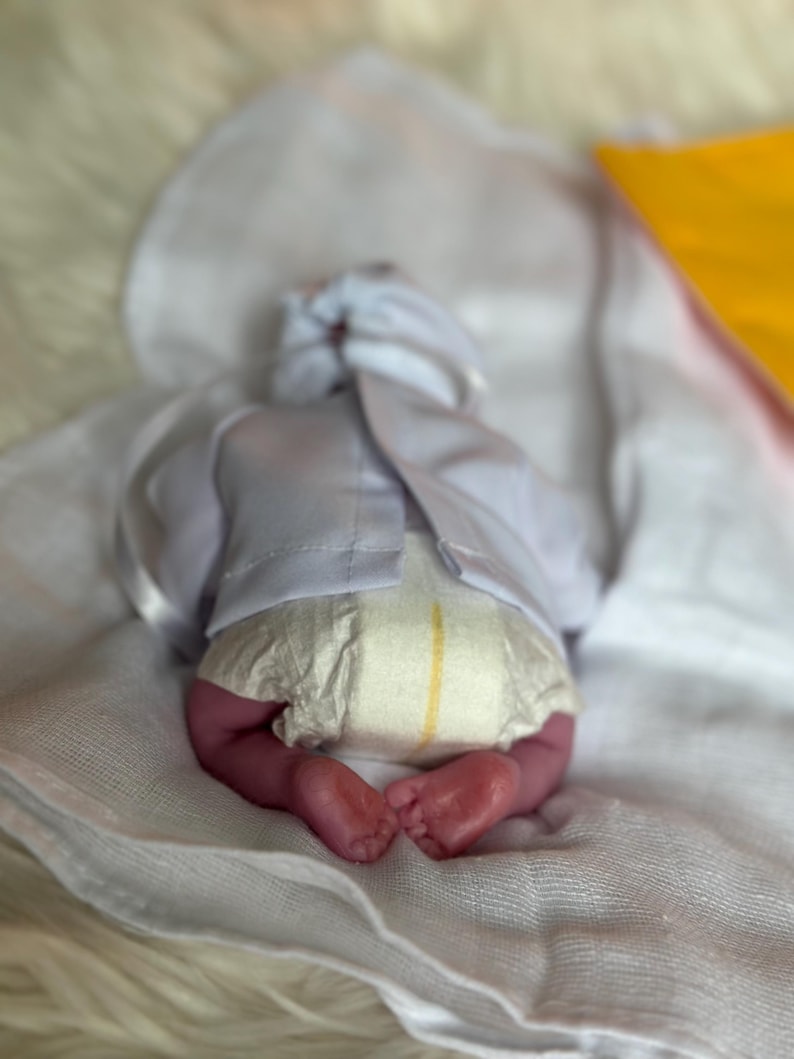 K&ouml;nnte beinhalten: Eine realistische Neugeborenenpuppe, die auf dem Bauch liegt und ein wei&szlig;es Langarmshirt mit R&uuml;ckenverschluss sowie eine Einwegwindel mit gelbem Feuchtigkeitsindikator tr&auml;gt. Die Puppe hat sichtbare Zehen und liegt auf einem wei&szlig;en Tuch.