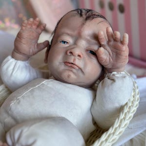 May include: A realistic reborn doll with pale skin, blue eyes, and sparse dark hair, wearing a long-sleeved cream-colored onesie. The doll is positioned in a white woven basket, with both hands raised near its face, showcasing detailed skin texture.