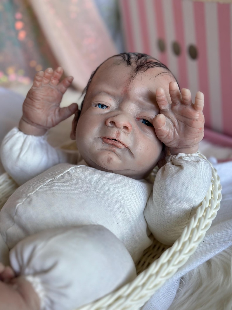 May include: A realistic reborn doll with pale skin, blue eyes, and sparse dark hair, wearing a long-sleeved cream-colored onesie. The doll is positioned in a white woven basket, with both hands raised near its face, showcasing detailed skin texture.