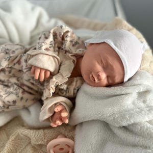 May include: A realistic newborn doll with closed eyes, wearing a white cap and a long-sleeved beige romper featuring a delicate floral pattern with ruffled details. A light pink pacifier is placed nearby on a soft, textured cream-colored blanket.