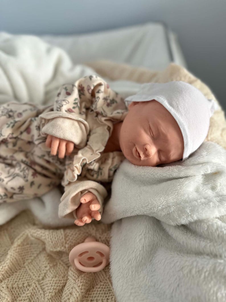 May include: A realistic newborn doll with closed eyes, wearing a white cap and a long-sleeved beige romper featuring a delicate floral pattern with ruffled details. A light pink pacifier is placed nearby on a soft, textured cream-colored blanket.