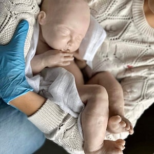 May include: A realistic baby doll with closed eyes, wearing a white cloth diaper, is being held by a person wearing a white sweater and blue jeans.