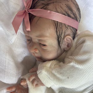 May include: A realistic reborn doll with dark, sparse hair and a light pink satin ribbon headband. The doll has fair skin with subtle veining, closed eyes, and is wearing a long-sleeved, white ribbed cotton shirt with a small button detail.