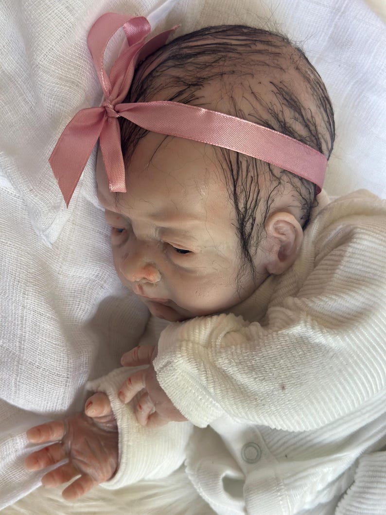 May include: A realistic reborn doll with dark, sparse hair and a light pink satin ribbon headband. The doll has fair skin with subtle veining, closed eyes, and is wearing a long-sleeved, white ribbed cotton shirt with a small button detail.