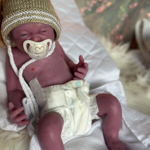Puede incluir: Una mu&ntilde;eca beb&eacute; realista con un chupete, que lleva un gorro de punto beige con una raya blanca. La mu&ntilde;eca tiene un pa&ntilde;al y est&aacute; tumbada sobre una superficie blanca. La piel de la mu&ntilde;eca tiene un tono viol&aacute;ceo.