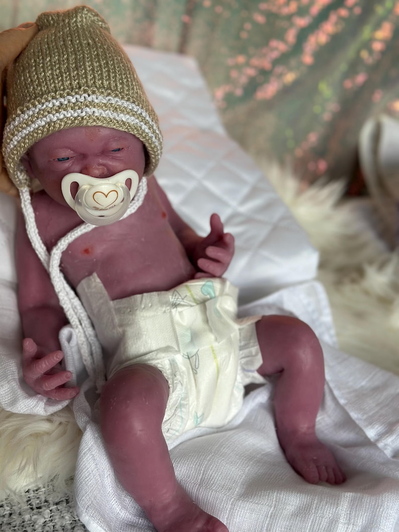 Puede incluir: Una mu&ntilde;eca beb&eacute; realista con un chupete, que lleva un gorro de punto beige con una raya blanca. La mu&ntilde;eca tiene un pa&ntilde;al y est&aacute; tumbada sobre una superficie blanca. La piel de la mu&ntilde;eca tiene un tono viol&aacute;ceo.