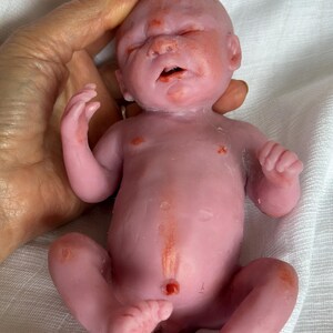 K&ouml;nnte beinhalten: Eine kleine, blassrosa Silikonpuppe in Form eines Neugeborenen mit geschlossenen Augen, leicht ge&ouml;ffnetem Mund und r&ouml;tlichen Hautdetails an Gesicht, Brust und Gliedma&szlig;en. Die Puppe verf&uuml;gt &uuml;ber einen realistischen Bauchnabel und Finger.