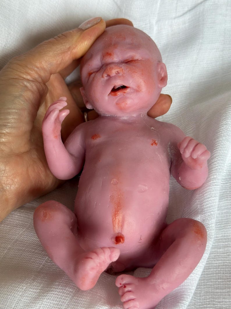 K&ouml;nnte beinhalten: Eine kleine, blassrosa Silikonpuppe in Form eines Neugeborenen mit geschlossenen Augen, leicht ge&ouml;ffnetem Mund und r&ouml;tlichen Hautdetails an Gesicht, Brust und Gliedma&szlig;en. Die Puppe verf&uuml;gt &uuml;ber einen realistischen Bauchnabel und Finger.