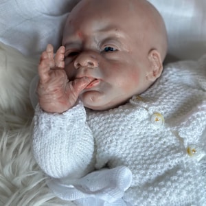 Może przedstawiać: Realistyczna lalka typu reborn o bladej, nakrapianej skórze, niebieskich oczach i łysej głowie. Lalka trzyma jedną dłoń przy ustach i ma na sobie biały, dzianinowy kardigan z długim rękawem o wyraźnej fakturze oraz małe, okrągłe guziki.