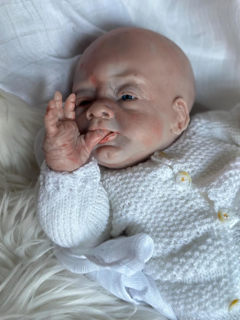 May include: A realistic reborn doll with pale, mottled skin, blue eyes, and a bald head. The doll has one hand raised to its mouth and is wearing a white, long-sleeved, textured knit cardigan featuring small, round, white buttons with gold centers.