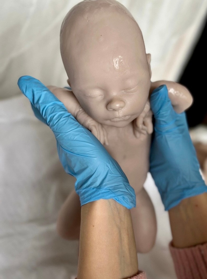 Puede incluir: Un mu&ntilde;eco de beb&eacute; de silicona realista con los ojos cerrados y la cara pintada. El mu&ntilde;eco est&aacute; siendo sostenido por manos enguantadas.
