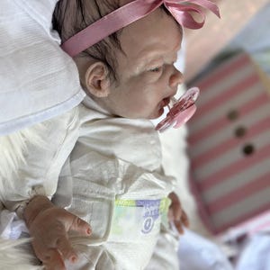 May include: A realistic newborn doll with dark hair, wearing a pink satin headband and a white long-sleeved onesie. The doll has a light skin tone, a pink pacifier in its mouth, and is wearing a disposable diaper with visible printed details.