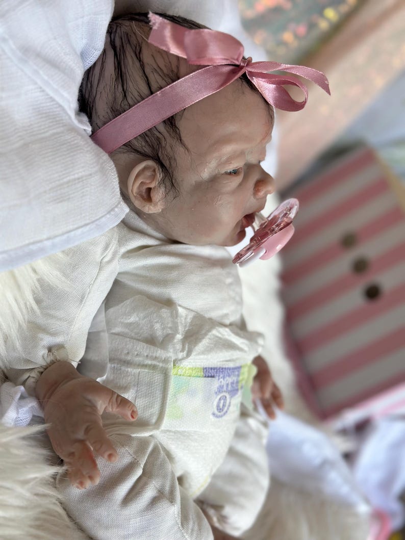 May include: A realistic newborn doll with dark hair, wearing a pink satin headband and a white long-sleeved onesie. The doll has a light skin tone, a pink pacifier in its mouth, and is wearing a disposable diaper with visible printed details.