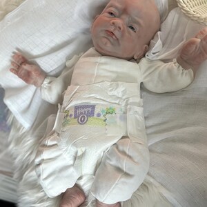May include: A realistic baby doll with pale skin, blue eyes, and a bald head, wearing a white long-sleeved onesie and a disposable diaper with the word Happy printed on the front. The doll is positioned on a white fabric surface.