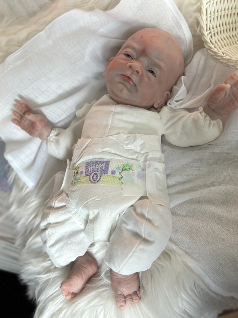 May include: A realistic baby doll with pale skin, blue eyes, and a bald head, wearing a white long-sleeved onesie and a disposable diaper with the word Happy printed on the front. The doll is positioned on a white fabric surface.