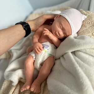 May include: A realistic newborn doll with light skin, closed eyes, and a subtle smile. The doll wears a plain white beanie cap and a disposable diaper with a colorful graphic print. It is resting on a soft, textured cream-colored fabric blanket.