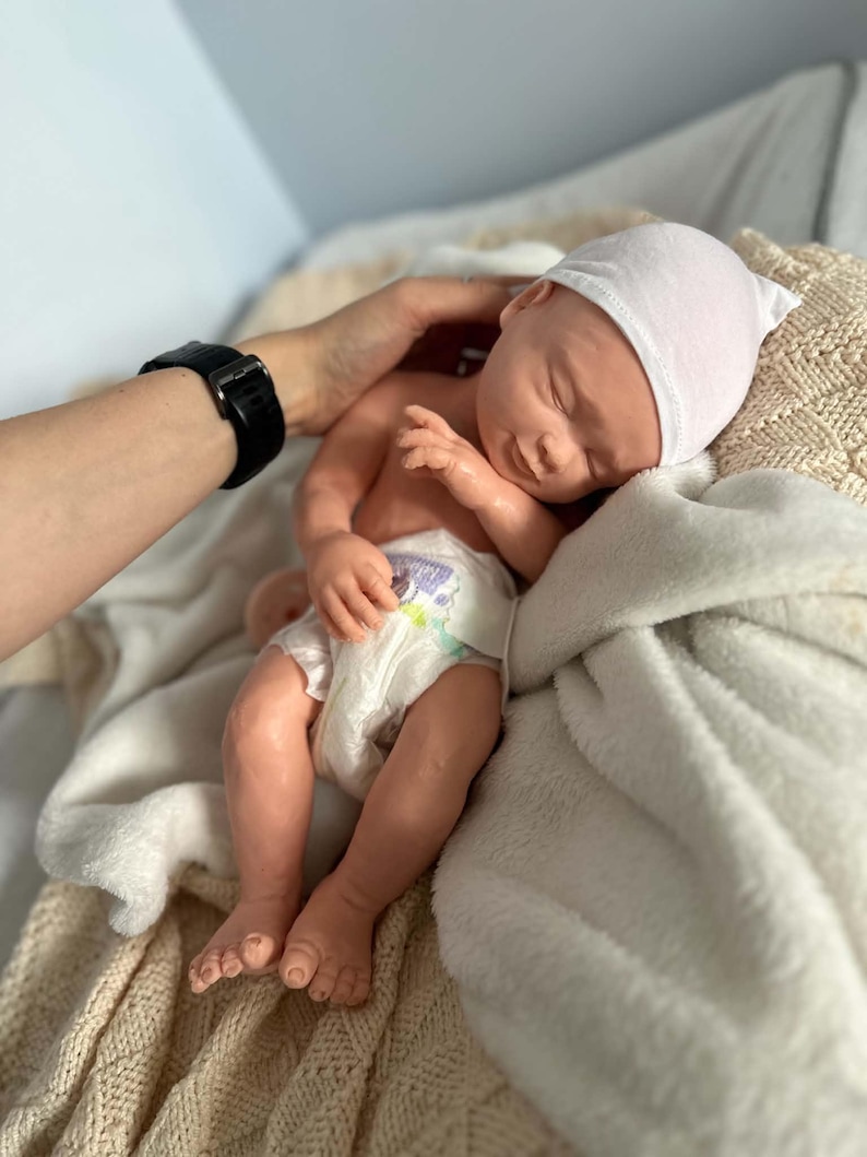 May include: A realistic newborn doll with light skin, closed eyes, and a subtle smile. The doll wears a plain white beanie cap and a disposable diaper with a colorful graphic print. It is resting on a soft, textured cream-colored fabric blanket.