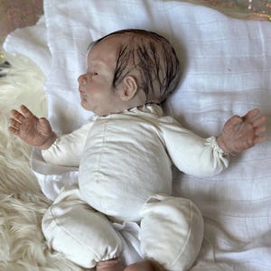 May include: A realistic newborn doll with painted dark hair, light skin, and detailed facial features. The doll is wearing a long-sleeved, off-white cotton onesie with gathered cuffs at the wrists and ankles, lying on a white muslin cloth.
