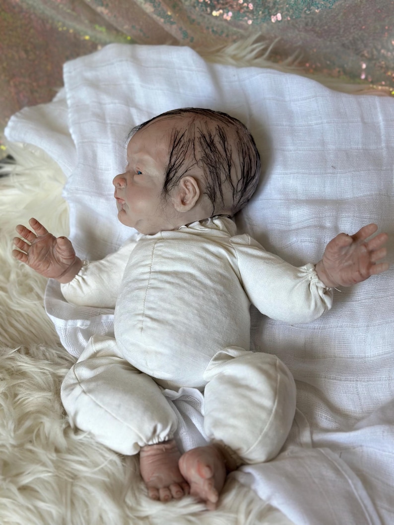 May include: A realistic newborn doll with painted dark hair, light skin, and detailed facial features. The doll is wearing a long-sleeved, off-white cotton onesie with gathered cuffs at the wrists and ankles, lying on a white muslin cloth.