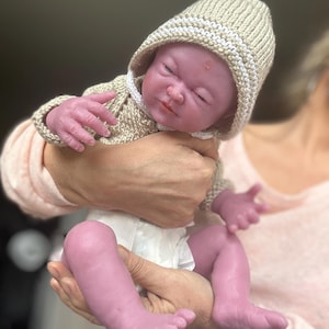 Puede incluir: Una mu&ntilde;eca beb&eacute; realista con los ojos cerrados, vestida con un su&eacute;ter con capucha de punto beige y un pa&ntilde;al. La piel de la mu&ntilde;eca tiene un tono viol&aacute;ceo. La mu&ntilde;eca est&aacute; en manos de alguien.