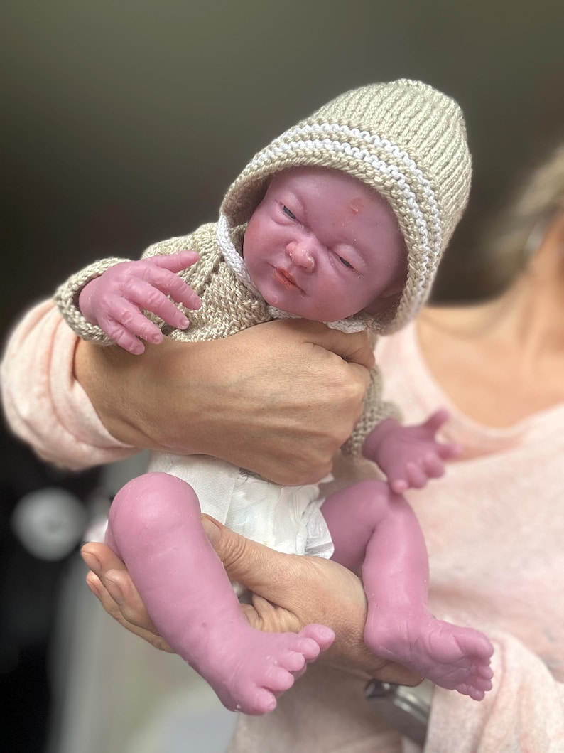 Puede incluir: Una mu&ntilde;eca beb&eacute; realista con los ojos cerrados, vestida con un su&eacute;ter con capucha de punto beige y un pa&ntilde;al. La piel de la mu&ntilde;eca tiene un tono viol&aacute;ceo. La mu&ntilde;eca est&aacute; en manos de alguien.