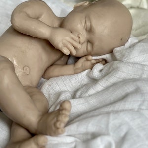 K&ouml;nnte beinhalten: Eine lebensechte Babypuppe mit nat&uuml;rlichem Hautton, die auf einer wei&szlig;en, strukturierten Decke schl&auml;ft. Die Hand der Puppe liegt in der N&auml;he ihres Gesichts, und ihre Augen sind geschlossen. Die F&uuml;&szlig;e des Babys sind ebenfalls sichtbar.