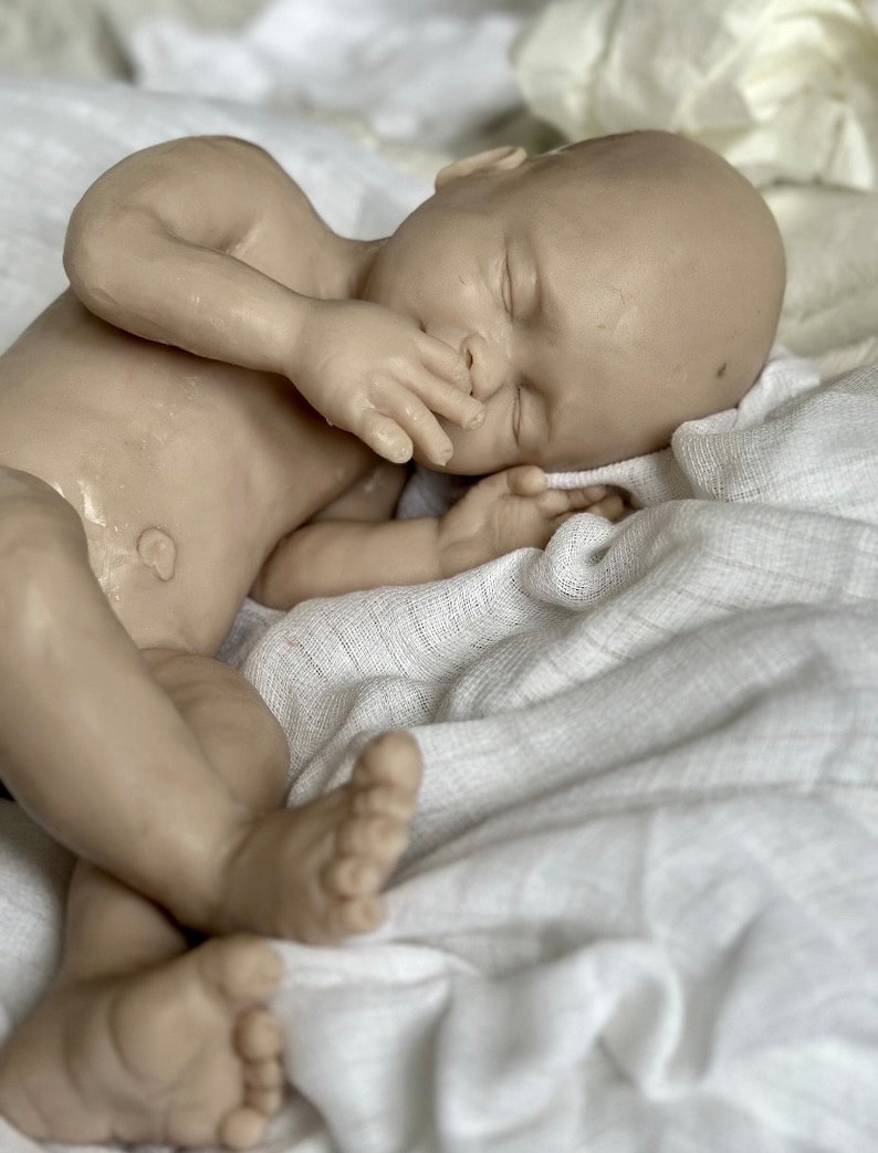 K&ouml;nnte beinhalten: Eine lebensechte Babypuppe mit nat&uuml;rlichem Hautton, die auf einer wei&szlig;en, strukturierten Decke schl&auml;ft. Die Hand der Puppe liegt in der N&auml;he ihres Gesichts, und ihre Augen sind geschlossen. Die F&uuml;&szlig;e des Babys sind ebenfalls sichtbar.