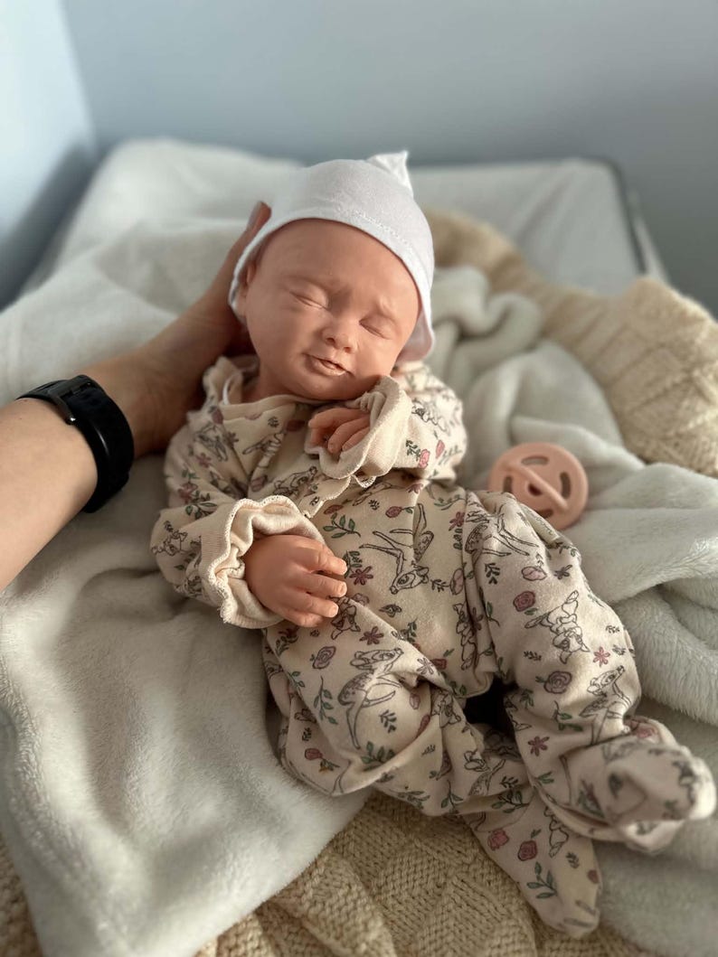 May include: A realistic baby doll with closed eyes and a neutral expression, wearing a white knit cap and a beige long-sleeved footed sleeper featuring a repeating pattern of small deer and floral motifs, accompanied by a round light pink pacifier.