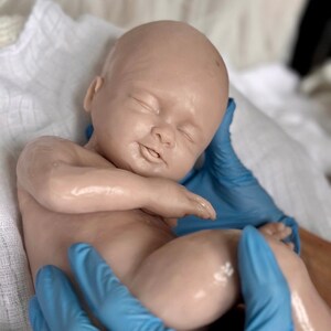 K&ouml;nnte beinhalten: Eine lebensechte Babypuppe mit heller Haut und geschlossenen Augen, die auf einem wei&szlig;en Tuch liegt. Die Puppe wird von H&auml;nden gehalten, die blaue Handschuhe tragen. Die Haut des Babys weist feine Details auf, und die Puppe wirkt entspannt.