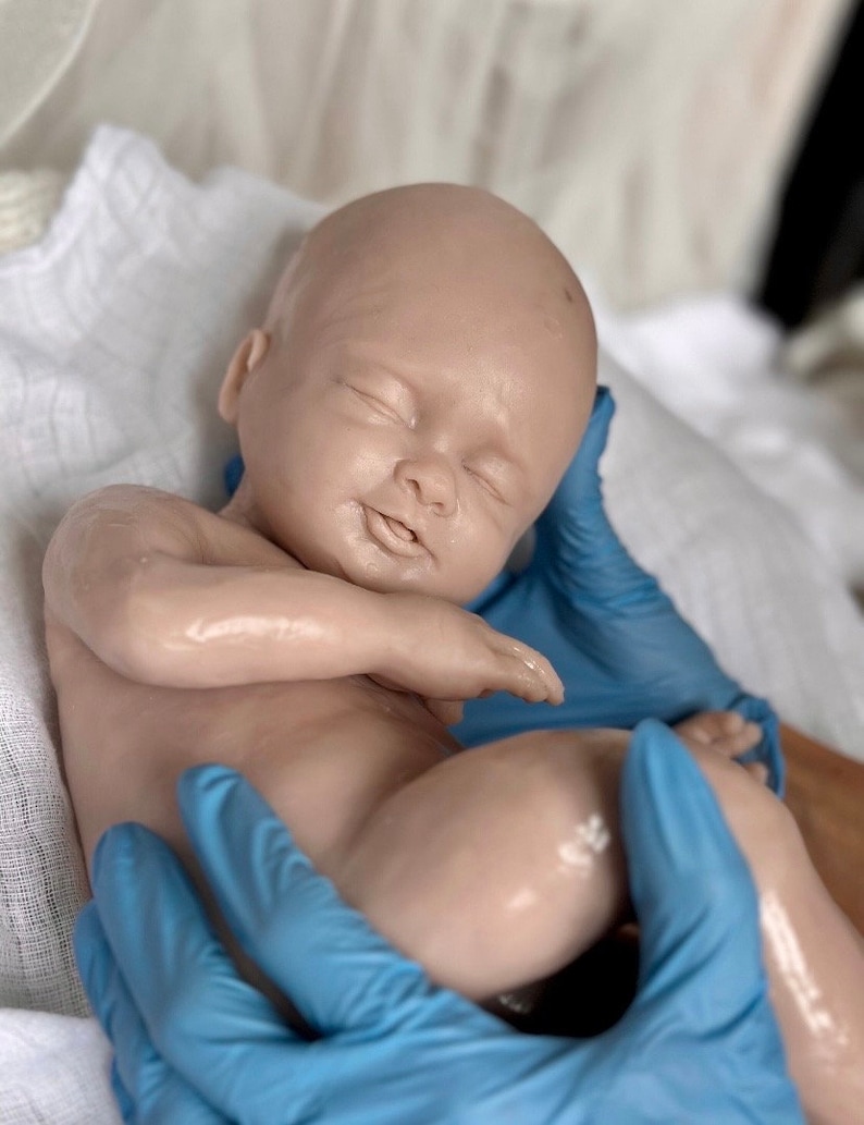 K&ouml;nnte beinhalten: Eine lebensechte Babypuppe mit heller Haut und geschlossenen Augen, die auf einem wei&szlig;en Tuch liegt. Die Puppe wird von H&auml;nden gehalten, die blaue Handschuhe tragen. Die Haut des Babys weist feine Details auf, und die Puppe wirkt entspannt.