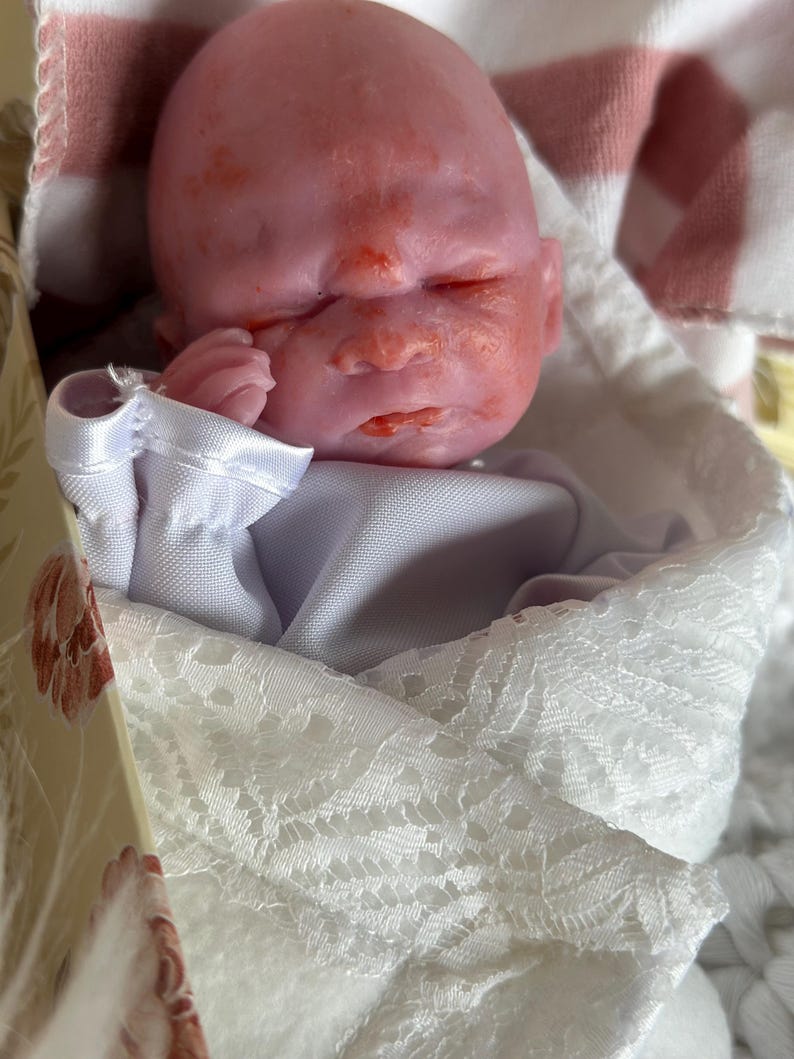 K&ouml;nnte beinhalten: Eine Miniatur-Silikonpuppe mit blassrosa Teint und r&ouml;tlicher Hautfleckung, geschlossenen Augen und einer kleinen Hand in Gesichtsn&auml;he. Die Puppe ist in ein wei&szlig;es Stoffgewand mit zartem Spitzenbesatz und einem wei&szlig;en Satin-Kragen eingewickelt.