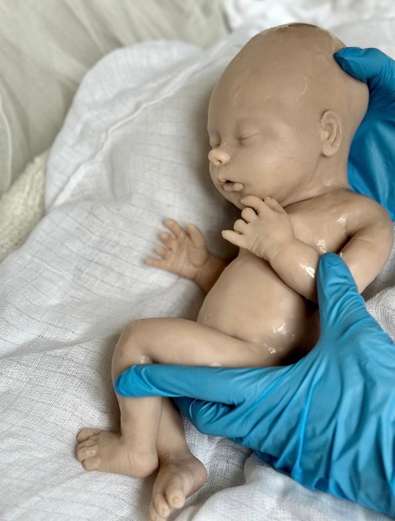 Puede incluir: Un mu&ntilde;eco de beb&eacute; realista hecho de silicona, acostado sobre una superficie de tela blanca. El mu&ntilde;eco lleva un guante azul en la pierna y otro guante azul sostiene el brazo del mu&ntilde;eco.