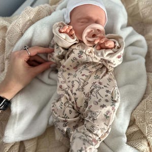 May include: A realistic baby doll wearing a cream-colored long-sleeve footed sleeper with a repeating pattern of small deer and floral motifs. The doll has a white beanie cap and a light pink pacifier in its mouth, resting on a soft white blanket.