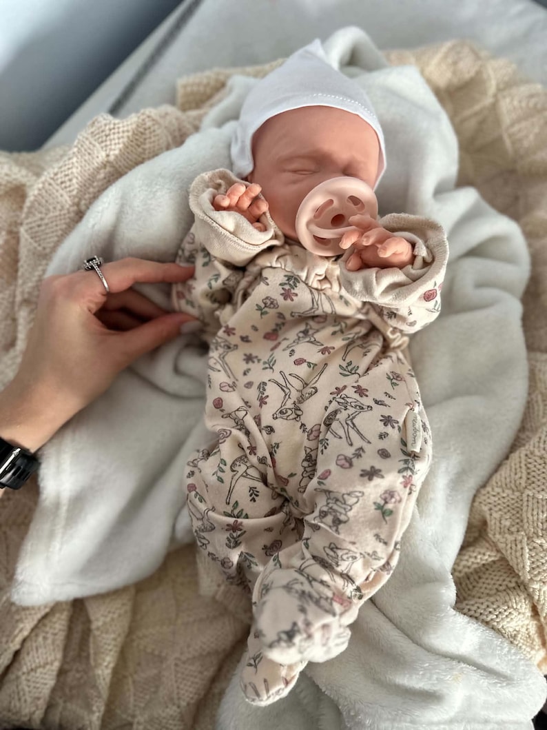 May include: A realistic baby doll wearing a cream-colored long-sleeve footed sleeper with a repeating pattern of small deer and floral motifs. The doll has a white beanie cap and a light pink pacifier in its mouth, resting on a soft white blanket.