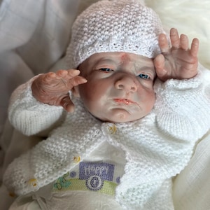 May include: A realistic reborn doll with pale skin, blue eyes, and a serious expression. The doll wears a white knitted hat and a matching white knitted cardigan with small yellow buttons over a white onesie featuring the word Happy in purple.