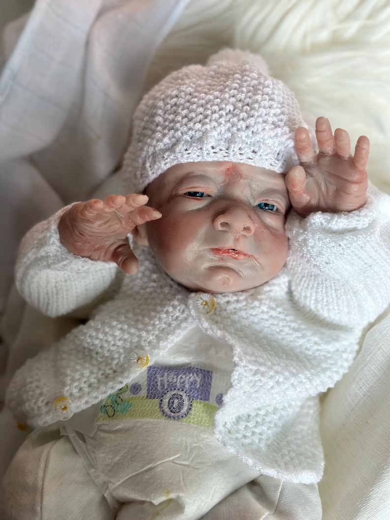 May include: A realistic reborn doll with pale skin, blue eyes, and a serious expression. The doll wears a white knitted hat and a matching white knitted cardigan with small yellow buttons over a white onesie featuring the word Happy in purple.
