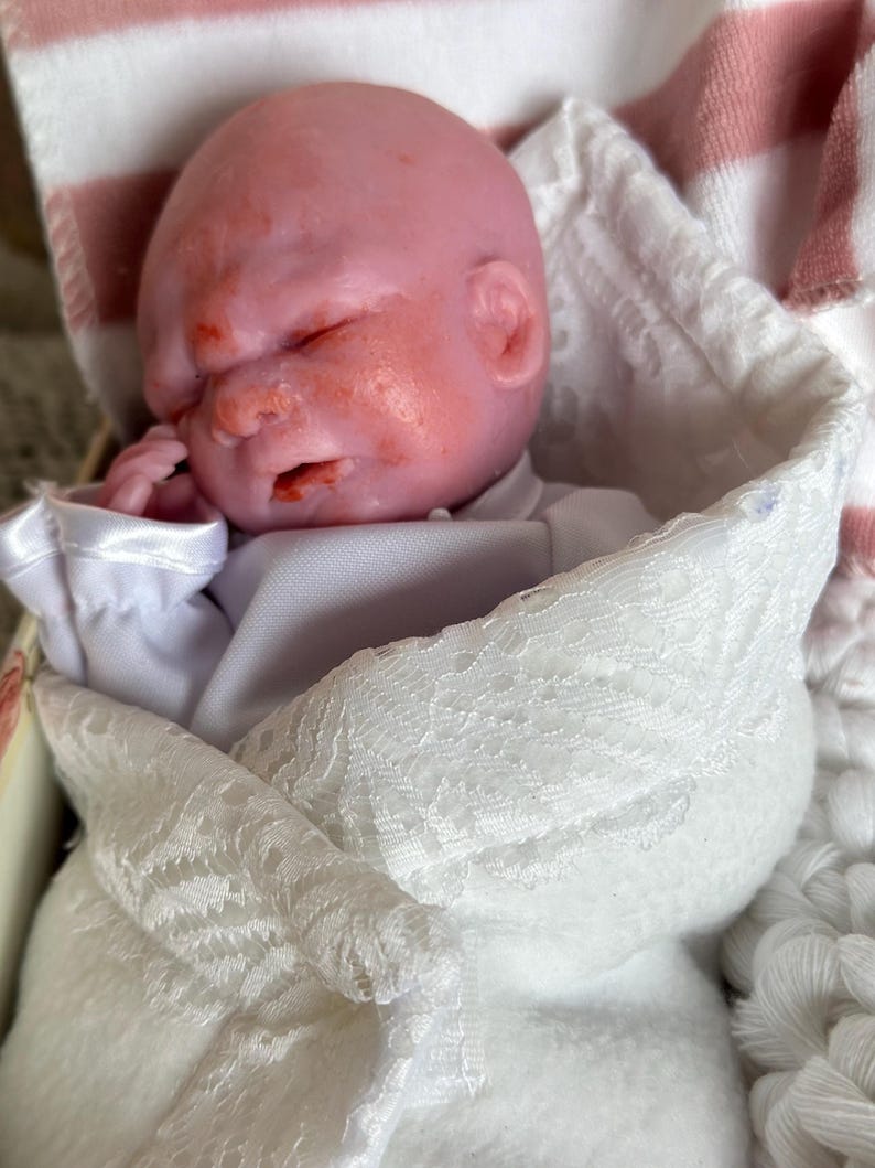 K&ouml;nnte beinhalten: Eine Miniaturpuppe mit blassrosa Teint und geschlossenen Augen, eingewickelt in ein wei&szlig;es Stofftuch mit zarter Spitzenborte. Die Puppe tr&auml;gt ein schlichtes wei&szlig;es Hemd und ist vor einem Hintergrund aus wei&szlig;-rosa gestreiftem Stoff platziert.