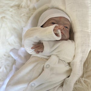 May include: A realistic newborn doll with pale skin and blue eyes, wearing a cream-colored ribbed long-sleeve onesie with snap buttons and a matching cap. The doll is wrapped in a white muslin cloth, showcasing detailed facial features and fingers.