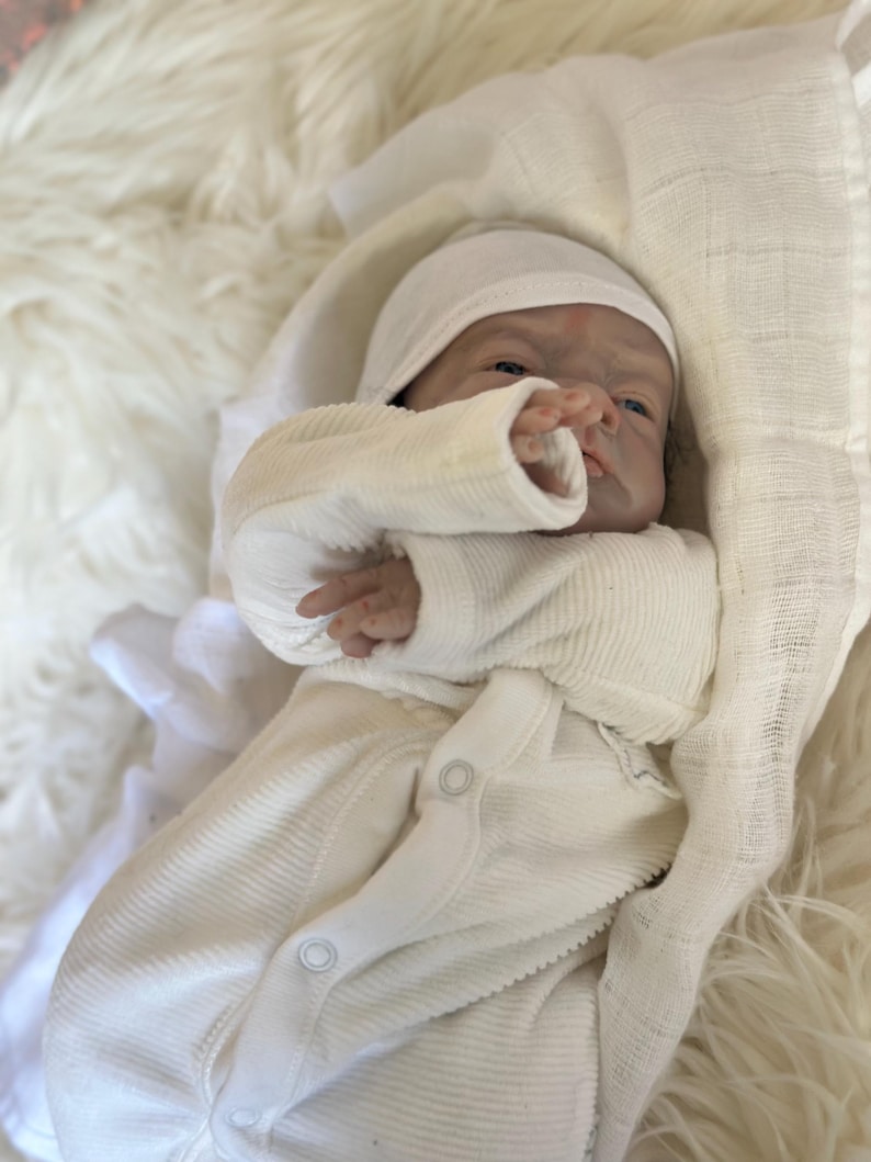 May include: A realistic newborn doll with pale skin and blue eyes, wearing a cream-colored ribbed long-sleeve onesie with snap buttons and a matching cap. The doll is wrapped in a white muslin cloth, showcasing detailed facial features and fingers.