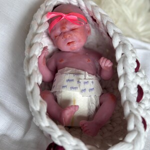 K&ouml;nnte beinhalten: Eine kleine, violett get&ouml;nte Silikonpuppe mit geschlossenen Augen und einem leuchtend rosa Schleifen-Stirnband, bekleidet mit einer wei&szlig;en Windel mit dem wiederholt aufgedruckten Wort Happy in Blau, in einem wei&szlig;en, ovalen H&auml;kelkorb.
