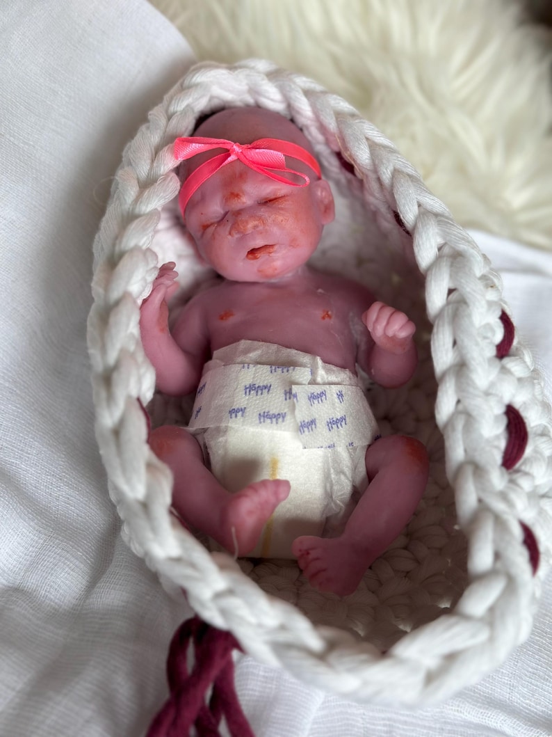 K&ouml;nnte beinhalten: Eine kleine, violett get&ouml;nte Silikonpuppe mit geschlossenen Augen und einem leuchtend rosa Schleifen-Stirnband, bekleidet mit einer wei&szlig;en Windel mit dem wiederholt aufgedruckten Wort Happy in Blau, in einem wei&szlig;en, ovalen H&auml;kelkorb.
