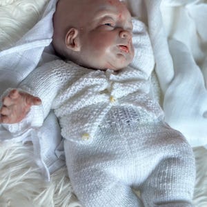 May include: A realistic reborn baby doll with pale skin, closed eyes, and subtle facial features, wearing a white knitted long-sleeve cardigan with small buttons and matching white knitted pants, lying down on a soft white textured fabric.
