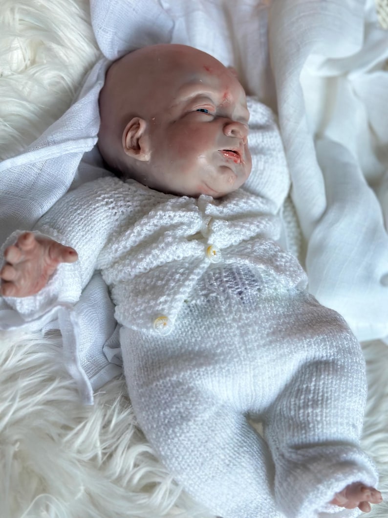 May include: A realistic reborn baby doll with pale skin, closed eyes, and subtle facial features, wearing a white knitted long-sleeve cardigan with small buttons and matching white knitted pants, lying down on a soft white textured fabric.