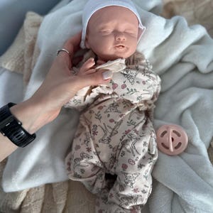 May include: A realistic newborn baby doll with closed eyes, wearing a white cap and a beige footed sleeper featuring a repeating pattern of small deer and pink flowers. A round, light pink pacifier is positioned next to the doll.