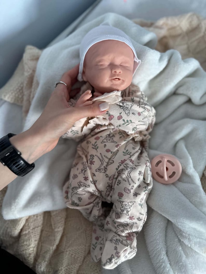 May include: A realistic newborn baby doll with closed eyes, wearing a white cap and a beige footed sleeper featuring a repeating pattern of small deer and pink flowers. A round, light pink pacifier is positioned next to the doll.