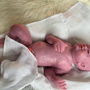 Puede incluir: Una mu&ntilde;eca beb&eacute; realista, de tono rosado, yace sobre una tela blanca. Los brazos y las piernas de la mu&ntilde;eca est&aacute;n ligeramente doblados, y tiene detalles realistas. Descansa sobre una superficie suave y blanquecina.