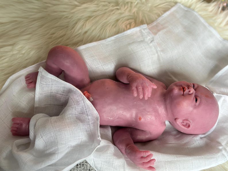 Puede incluir: Una mu&ntilde;eca beb&eacute; realista, de tono rosado, yace sobre una tela blanca. Los brazos y las piernas de la mu&ntilde;eca est&aacute;n ligeramente doblados, y tiene detalles realistas. Descansa sobre una superficie suave y blanquecina.