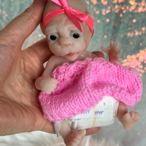 May include: A miniature doll with a pale complexion, wearing a pink knitted blanket and a matching headband with a bow. The doll is also wearing a nappy. The doll is held in a hand.