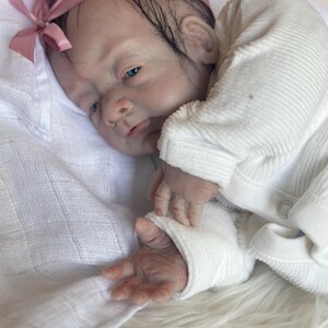 May include: A realistic reborn doll with pale skin, blue eyes, and dark hair, wearing a white long-sleeved ribbed shirt and a pink satin headband with a bow. The doll is resting on a white fabric surface, showcasing detailed facial features.